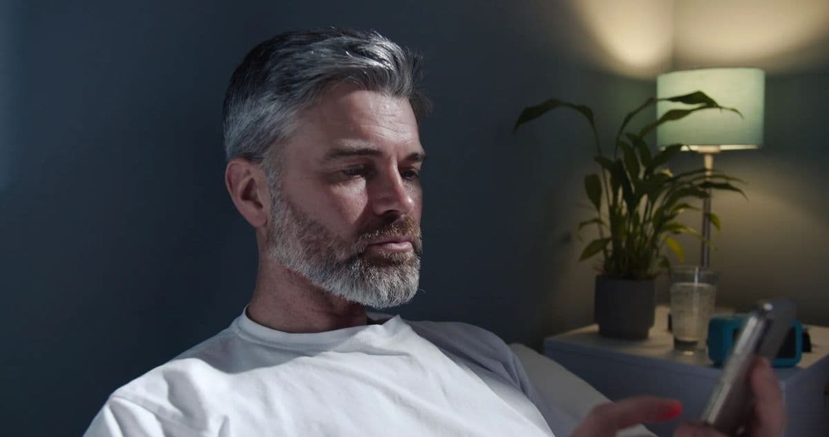 A middle-aged man staring at his phone while tired in his bedroom