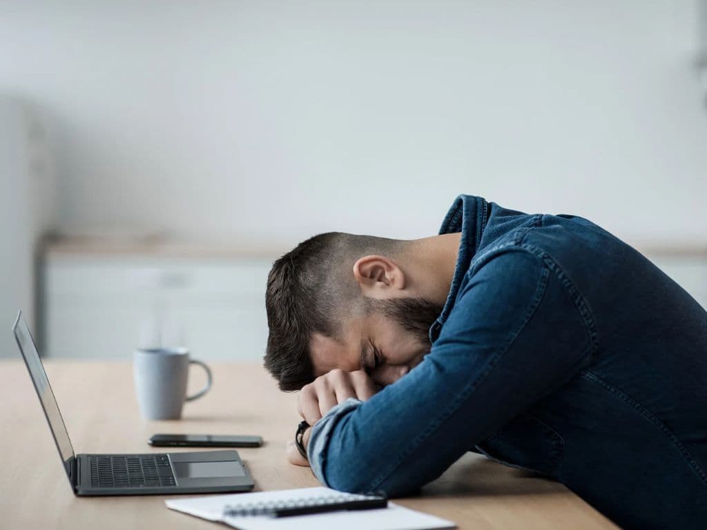 A man resting his head on his arms on his desk in front of his laptop